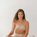 Full body of fit barefoot female with long hair wearing beige activewear looking at camera while sitting in lotus posture against white background